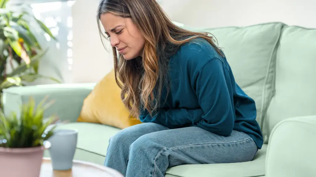 Woman sitting on a sofa and holding her lower abdomen while experiencing menstrual pain.